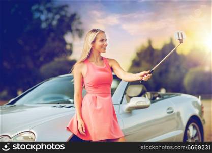 travel, road trip and people concept - happy young woman at convertible car taking picture by smartphone selfie stick over summer sunset background. woman taking picture by selfie stick at car. woman taking picture by selfie stick at car