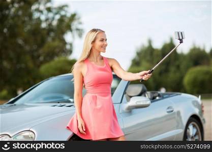 travel, road trip and people concept - happy young woman posing at convertible car and taking picture by smartphone selfie stick. woman taking picture by selfie stick at car