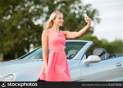 travel, road trip and people concept - happy young woman posing at convertible car and taking selfie by smartphone. woman posing at convertible car and taking selfie