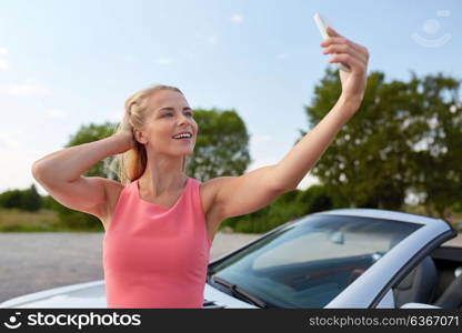 travel, road trip and people concept - happy young woman posing at convertible car and taking selfie by smartphone. woman posing at convertible car and taking selfie