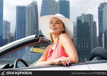 travel, road trip and people concept - happy young woman wearing sun hat in convertible car over singapore city skyscrapers background. woman in hat in convertible car over singapore