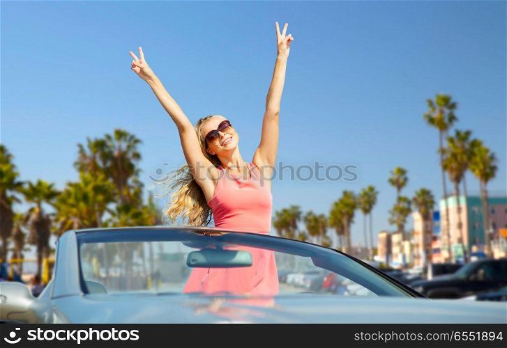 travel, summer holidays, road trip and people concept - happy young woman wearing sunglasses in convertible car showing peace hand sign over venice beach background in california. happy woman in convertible car over venice beach. happy woman in convertible car over venice beach