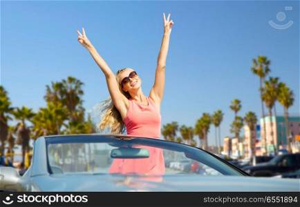 travel, summer holidays, road trip and people concept - happy young woman wearing sunglasses in convertible car showing peace hand sign over venice beach background in california. happy woman in convertible car over venice beach. happy woman in convertible car over venice beach