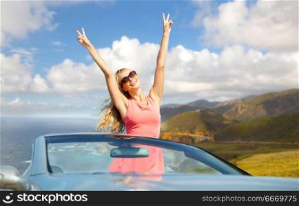 travel, summer holidays, road trip and people concept - happy young woman wearing sunglasses in convertible car showing peace sign over bixby creek bridge on big sur coast of california background. happy woman in convertible car on big sur coast. happy woman in convertible car on big sur coast