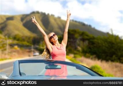 travel, summer holidays, road trip and people concept - happy young woman wearing sunglasses in convertible car showing peace hand sign over big sur hills and road background in california. happy young woman in convertible car. happy young woman in convertible car