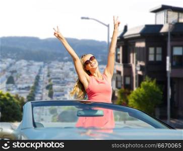 travel, summer holidays, road trip and people concept - happy young woman wearing sunglasses in convertible car showing peace hand sign over san francisco city background. happy woman in convertible car over san francisco. happy woman in convertible car over san francisco