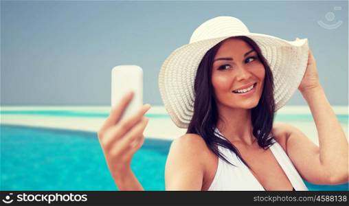 travel, summer, technology and people concept - sexy young woman taking selfie with smartphone over beach and swimming pool background