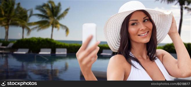travel, summer, technology and people concept - sexy young woman taking selfie with smartphone over resort beach with palms and swimming pool background