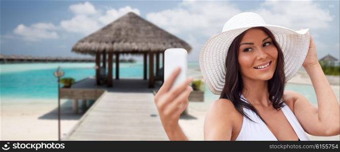 travel, summer, technology and people concept - sexy young woman taking selfie with smartphone over bungalow on beach background