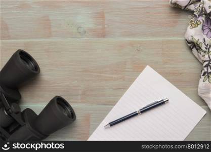 Travel, summer vacation, tourism and objects concept - close up binoculars, pen, paper and scarf on wooden table. Top view with copy space.