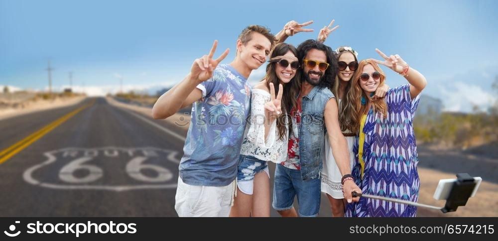 travel, technology and road trip concept - smiling young hippie friends taking picture by smartphone on selfie stick and showing peace hand sign over us route 66 background. hippie friends taking selfie over us route 66