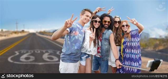 travel, technology and road trip concept - smiling young hippie friends taking picture by smartphone on selfie stick and showing peace hand sign over us route 66 background. hippie friends taking selfie over us route 66