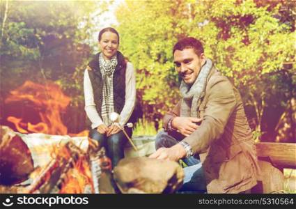 travel, tourism and hike concept - happy couple roasting marshmallow over campfire at camp in woods. happy couple roasting marshmallow over camp fire