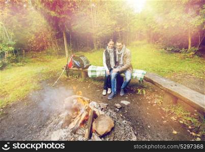 travel, tourism and hike concept - happy couple sitting on bench and warming near campfire at camp in woods. happy couple sitting on bench near camp fire