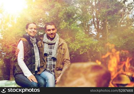 travel, tourism and hike concept - happy couple sitting on bench and warming near campfire at camp in woods. happy couple sitting on bench near camp fire
