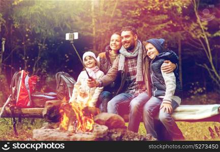 travel, tourism and hike concept - happy family sitting on bench and taking picture by smartphone on selfie stick at campfire in woods. family with smartphone taking selfie near campfire