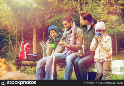 travel, tourism and hike concept - happy family sitting on bench and drinking hot tea from cups at camp fire in woods. happy family sitting on bench at camp fire