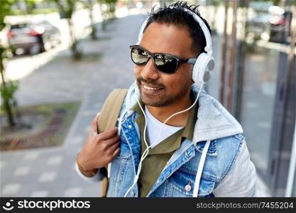travel, tourism and lifestyle concept - smiling indian man in headphones with backpack listening to music on city street. indian man in headphones with backpack in city