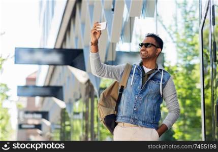travel, tourism and lifestyle concept - smiling indian man in sunglasses with backpack taking selfie by smartphone on city street. indian man taking selfie by smartphone in city