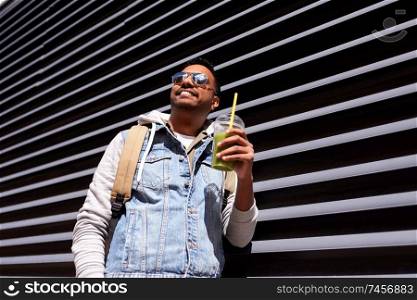 travel, tourism and lifestyle concept - smiling indian man with backpack drinking smoothie from plastic cup with straw on city street. man with backpack drinking smoothie on street