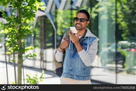 travel, tourism and lifestyle concept - smiling indian man with backpack using voice command recorder on smartphone on city street. man recorving voice message on smartphone in city