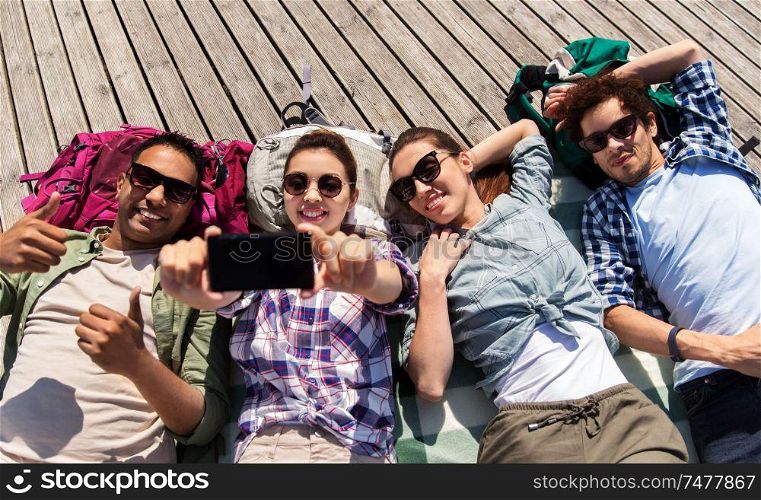 travel, tourism and people concept - group of friends or tourists with backpacks lying on wooden terrace and taking selfie by smartphone. friends or tourists with backpacks taking selfie