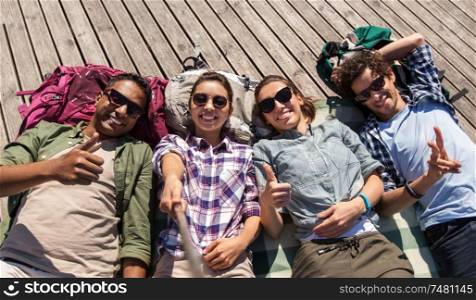 travel, tourism and people concept - group of friends or tourists with backpacks lying on wooden terrace and taking picture by selfie stick. friends or tourists with backpacks taking selfie