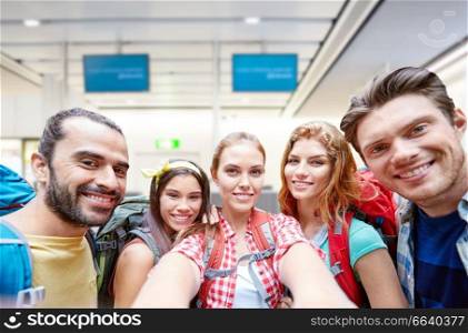 travel, tourism and people concept - group of smiling friends or tourists with backpacks taking selfie over airport terminal background. friends or tourists taking selfie over airport