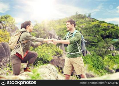 travel, tourism and people concept - happy couple with backpacks climbing over fallen tree trunk over exotic island hills background. couple with backpacks traveling over exotic island. couple with backpacks traveling over exotic island