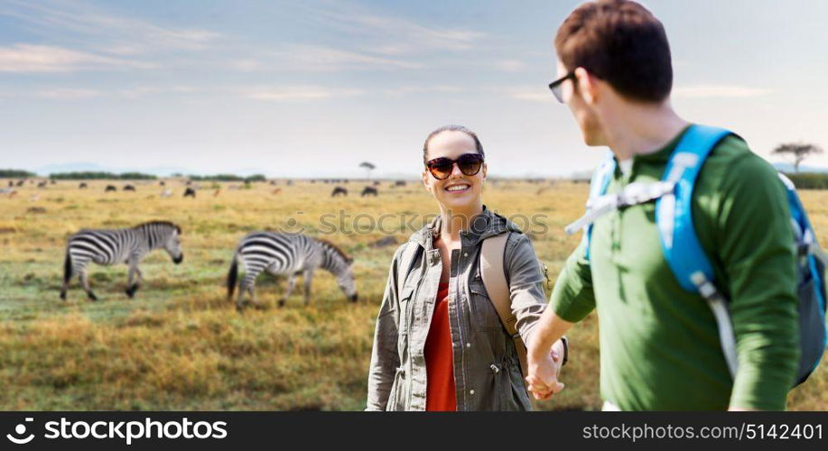 travel, tourism and people concept - happy couple with backpacks holding hands over african savannah and zebras background. smiling couple with backpacks traveling in africa