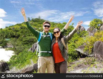 travel, tourism and people concept - happy couple with backpacks waving hands over natural island background. couple with backpacks traveling around island. couple with backpacks traveling around island