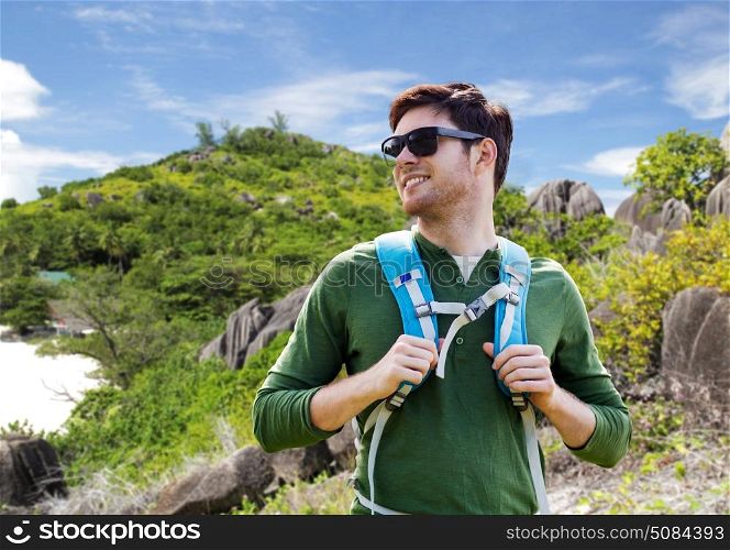 travel, tourism and people concept - happy young man in sunglasses with backpack over natural exotic island background. happy man with backpack traveling around island. happy man with backpack traveling around island