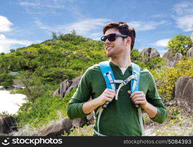 travel, tourism and people concept - happy young man in sunglasses with backpack over natural exotic island background. happy man with backpack traveling around island. happy man with backpack traveling around island