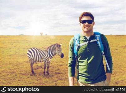 travel, tourism and people concept - happy young man in sunglasses with backpack over african savannah and zebra background. happy young man with backpack traveling in africa