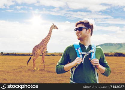 travel, tourism and people concept - happy young man in sunglasses with backpack over giraffe in african savannah background. happy man with backpack traveling in africa