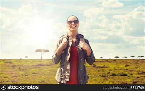 travel, tourism and people concept - happy young woman in sunglasses with backpack over african savannah background. happy woman with backpack traveling in africa. happy woman with backpack traveling in africa