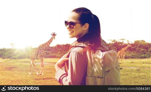 travel, tourism and people concept - happy young woman with backpack over african savannah and giraffe background. happy woman with backpack traveling in africa. happy woman with backpack traveling in africa