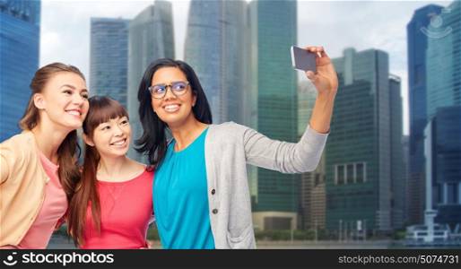 travel, tourism and people concept - international group of happy smiling different women taking selfie with smartphone over singapore city skyscrapers background. international happy women taking selfie in city