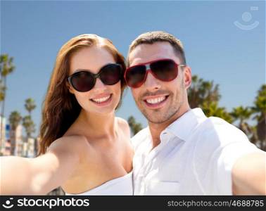travel, tourism and people concept - smiling couple wearing sunglasses making selfie over venice beach background in california. couple in shades making selfie over venice beach