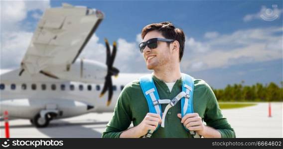 travel, tourism and people concept - smiling young man in sunglasses with backpack over plane on airfield background. smiling man with backpack over plane on airfield