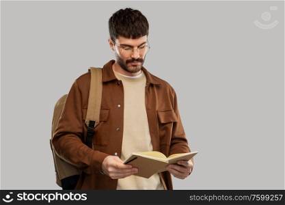 travel, tourism and people concept - young man in glasses with backpack reading diary over grey background. young man with backpack reading diary