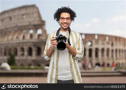 travel, tourism and photography concept - happy smiling man or photographer in glasses with digital camera over coliseum in rome, italy on background. smiling man or photographer with camera in rome