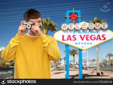 travel, tourism and photography concept - happy young male photographer in yellow sweatshirt taking picture with vintage film camera over welcome to fabulous las vegas sign background. man with vintage film camera over las vegas sign