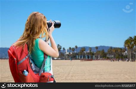 travel, tourism and photography concept - happy young woman with backpack and camera photographing over venice beach background in california. woman with backpack and camera over venice beach