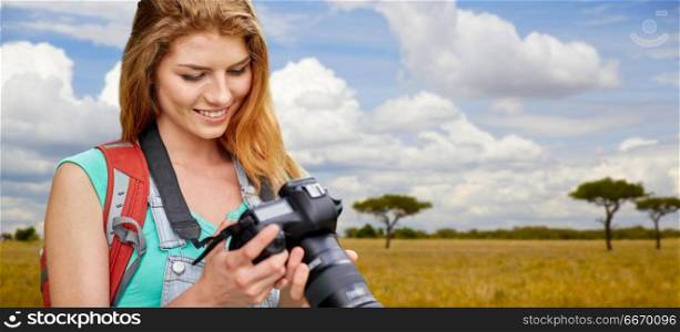 travel, tourism and photography concept - happy young woman with backpack and camera photographing over african savannah background. woman with backpack and camera over savannah. woman with backpack and camera over savannah