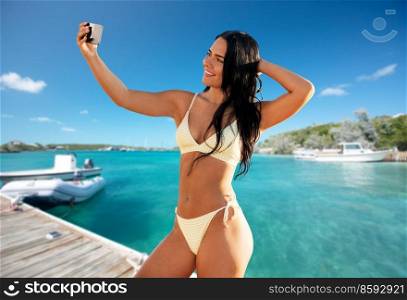 travel, tourism and summer concept - happy smiling young woman in bikini swimsuit taking selfie with smartphone over tropical beach background in french polynesia. smiling woman in bikini taking selfie on beach