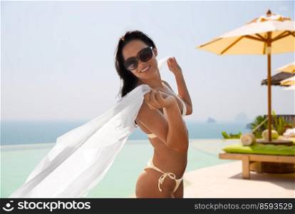 travel, tourism and summer concept - happy smiling young woman in bikini swimsuit with pareo over infinity edge swimming pool background. woman in bikini with pareo over infinity edge pool