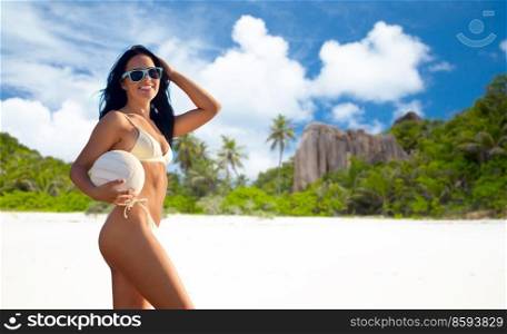 travel, tourism and summer concept - happy smiling young woman in bikini swimsuit posing with volleyball over seychelles island beach background. woman in bikini posing with volleyball on beach