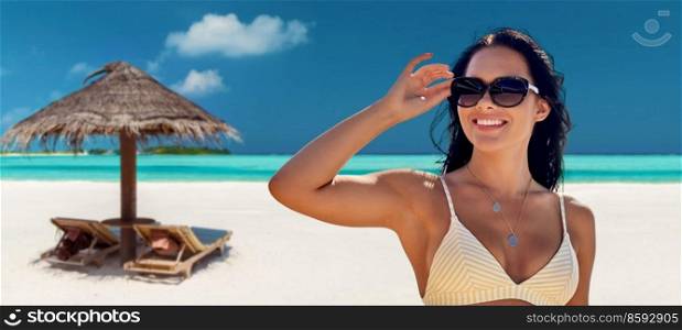 travel, tourism and summer concept - happy smiling young woman in sunglasses and bikini swimsuit over palapa and sun beds on maldives beach background. smiling young woman in sunglasses on summer beach