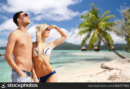 travel, tourism and summer holidays concept - happy couple in sunglasses at touristic resort over exotic tropical beach with palm tree and bungalow sheds background. happy couple over tropical beach. happy couple over tropical beach